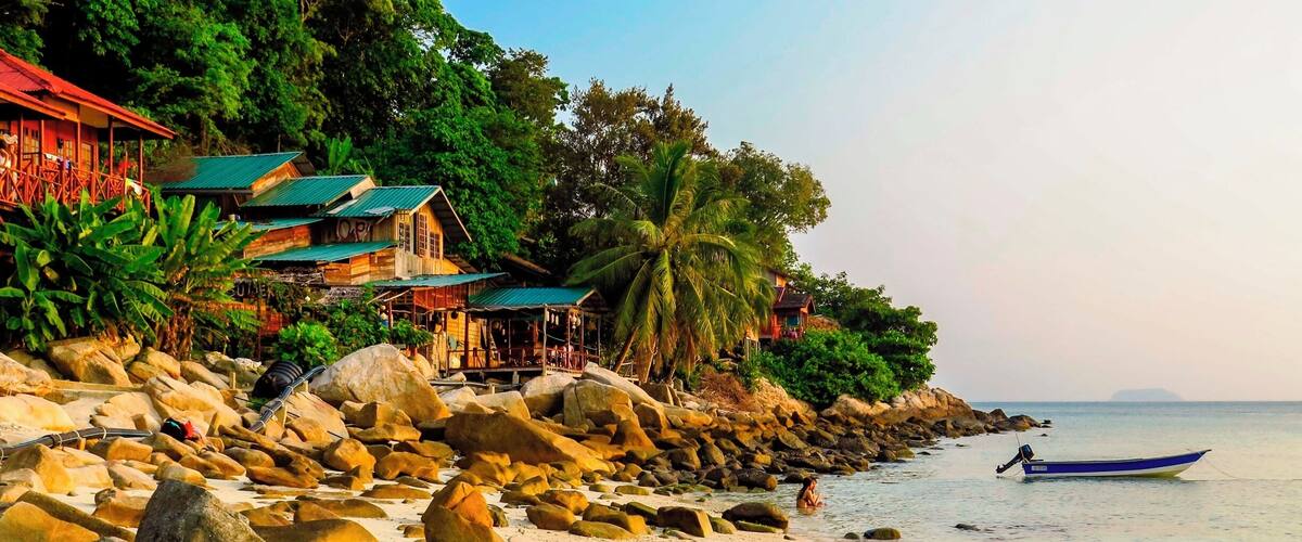 Une plage rocheuse parsemée de gros rochers, un petit bateau et une rangée de maisons aux toits verts.
