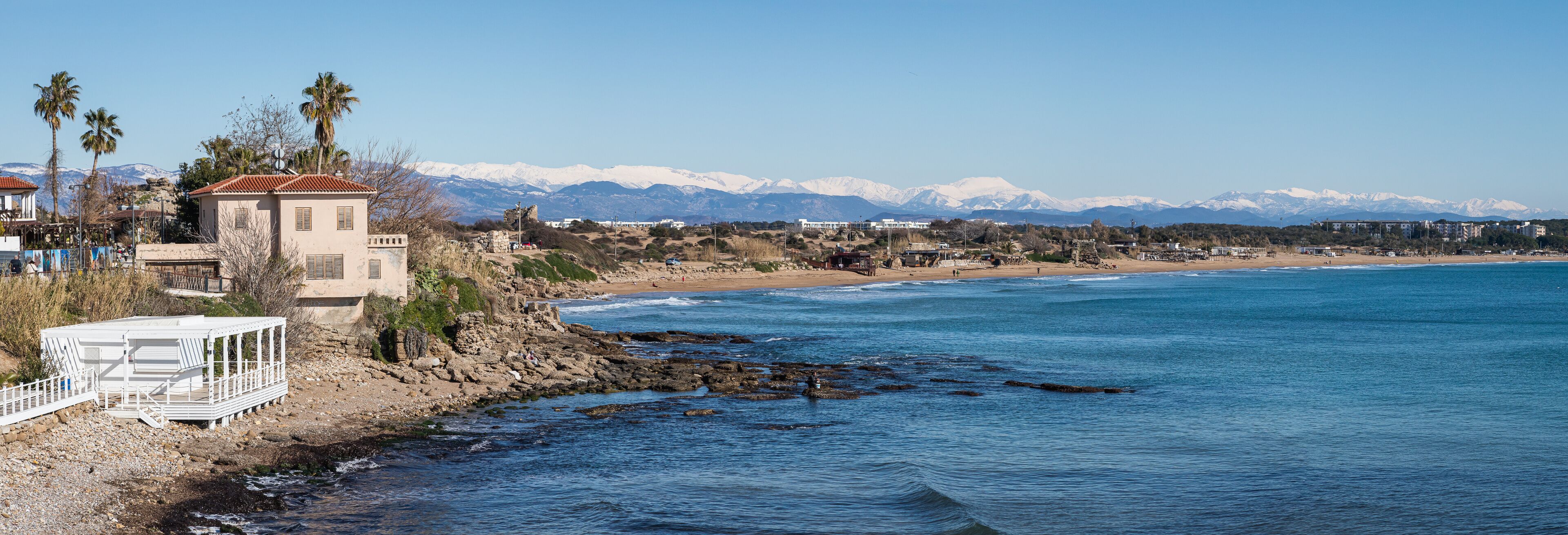 A coastal town with a rocky shoreline, a white pier, and snow-capped mountains in the background.