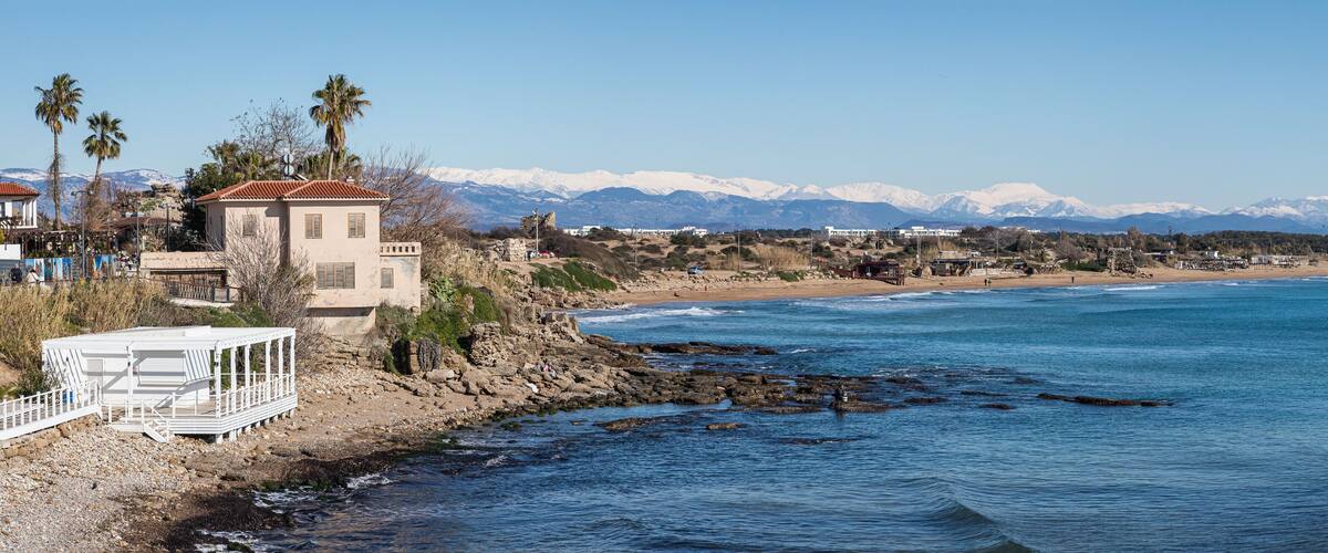 A coastal town with a rocky shoreline, a white pier, and snow-capped mountains in the background.