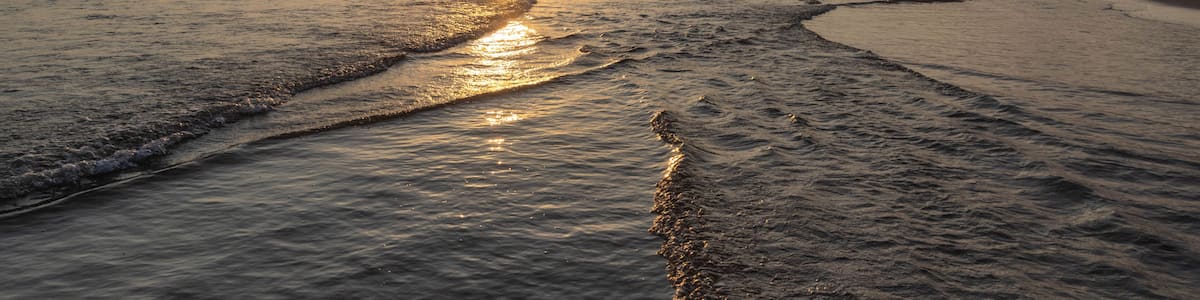 Sunset over shallow sandy beach with layered waves creating sparkling patterns, people silhouettes in sea, and parasailing canopy against golden sky. Evrenseki, Antalya, Turkey.
