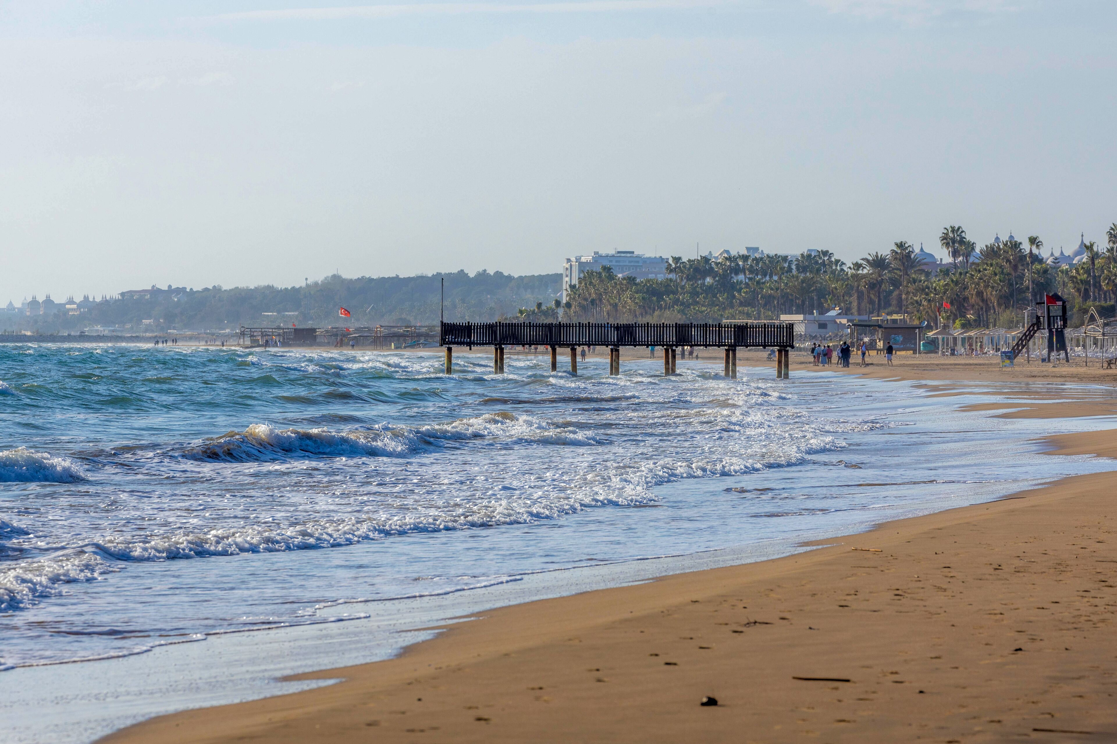 Telephoto of Evrenseki’s curved beach: foamy waves crash on the left, while a sandy cove with strolling visitors nestles under palm-fringed hotels. Manavgat, Turkey. March.