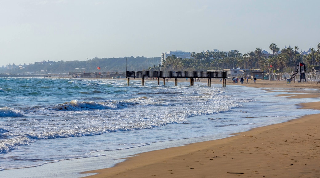 Telephoto of Evrenseki’s curved beach: foamy waves crash on the left, while a sandy cove with strolling visitors nestles under palm-fringed hotels. Manavgat, Turkey. March.