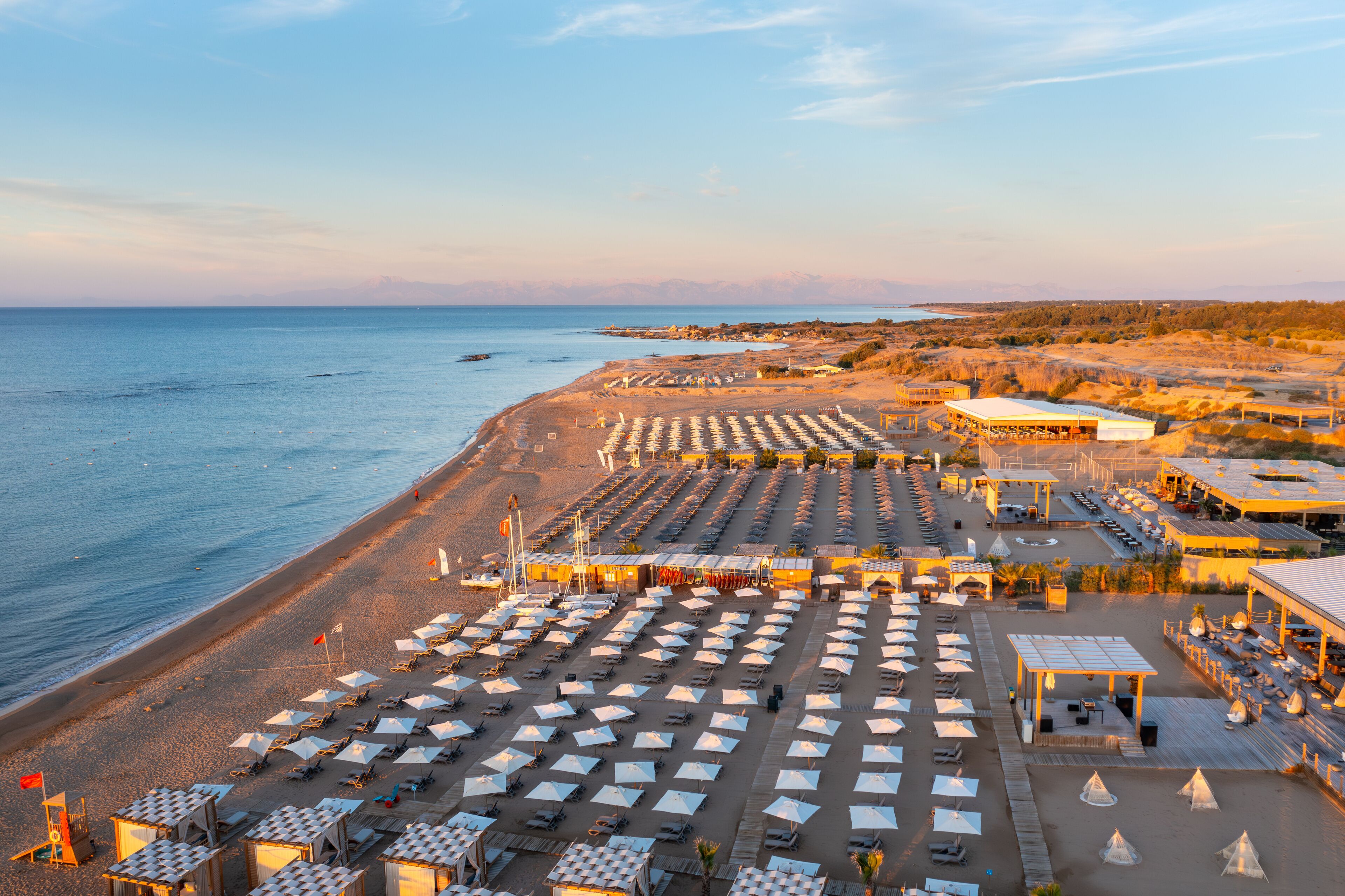 A beach with rows of umbrellas and sun loungers, a lifeguard tower, and a distant mountain range.