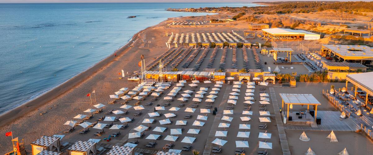A beach with rows of umbrellas and sun loungers, a lifeguard tower, and a distant mountain range.
