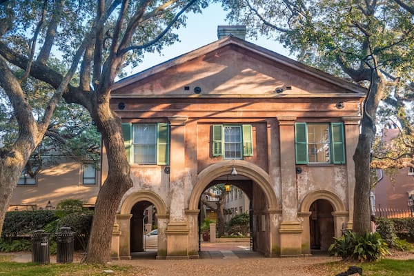 A historic building with green shutters, a central archway, and mature trees in the foreground.