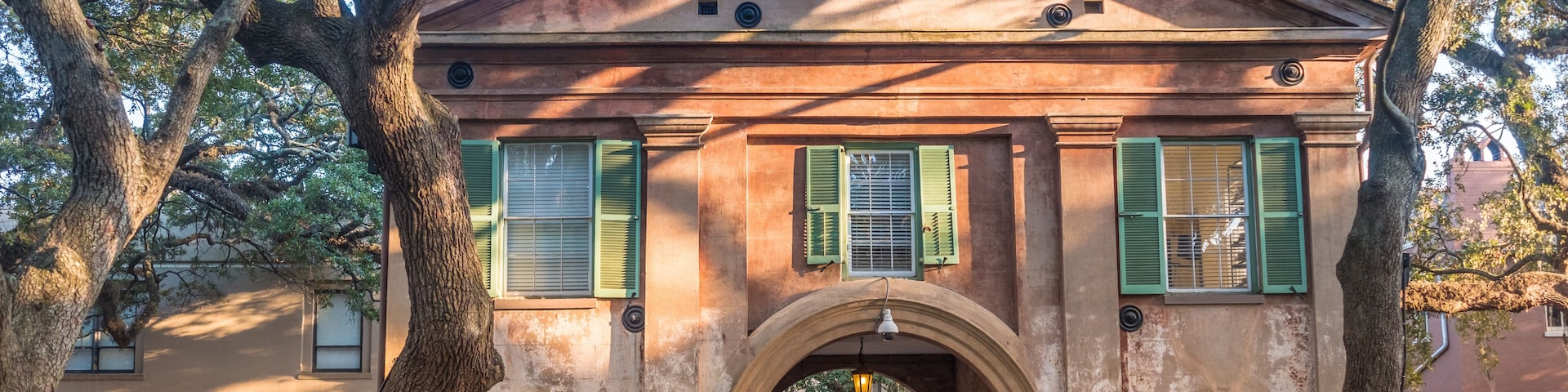 Um edifício histórico com portadas verdes, um arco central e árvores grandes em primeiro plano.