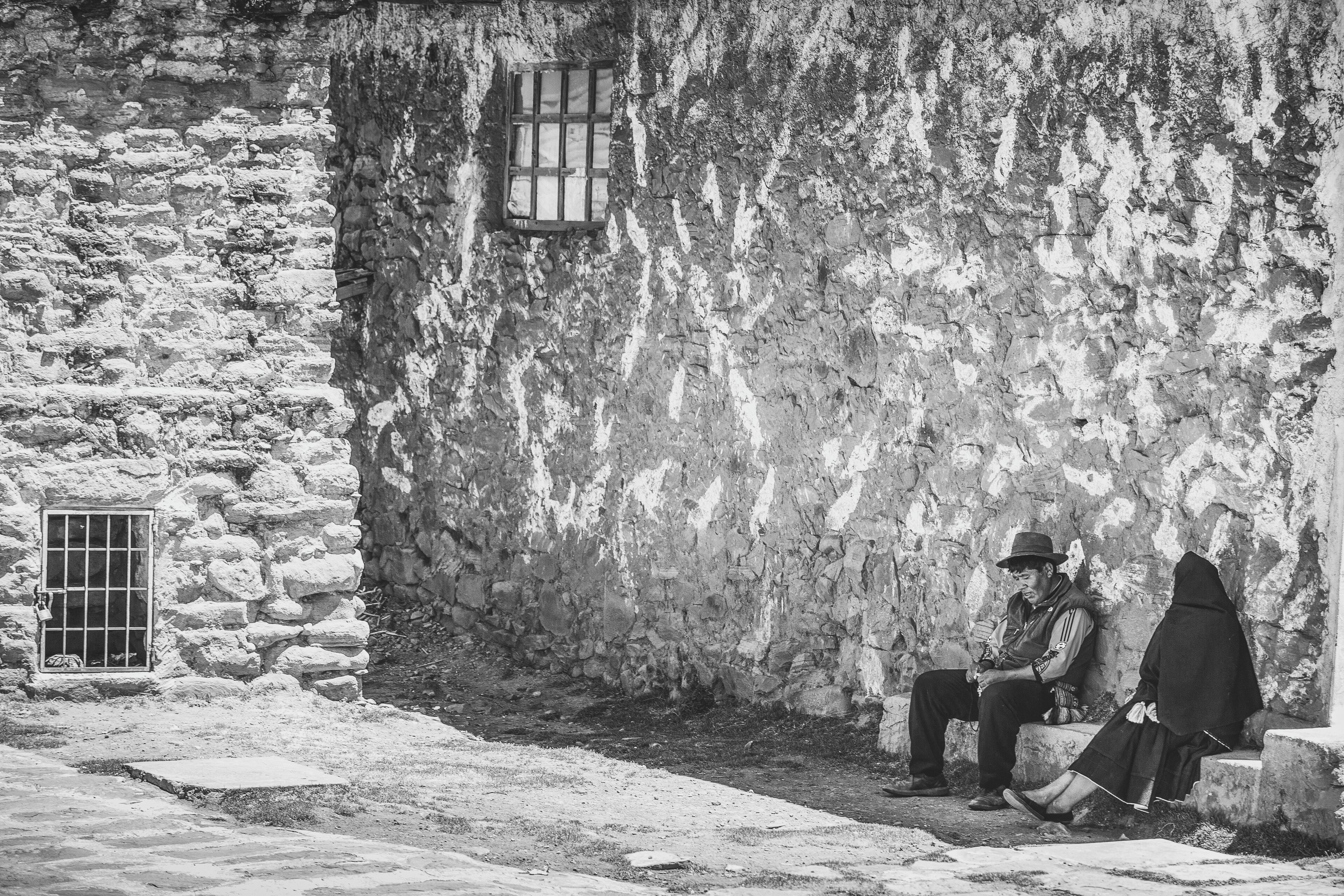 Un edificio de piedra con una ventana y una puerta, una persona sentada en un banco y otra con una capa con capucha.
