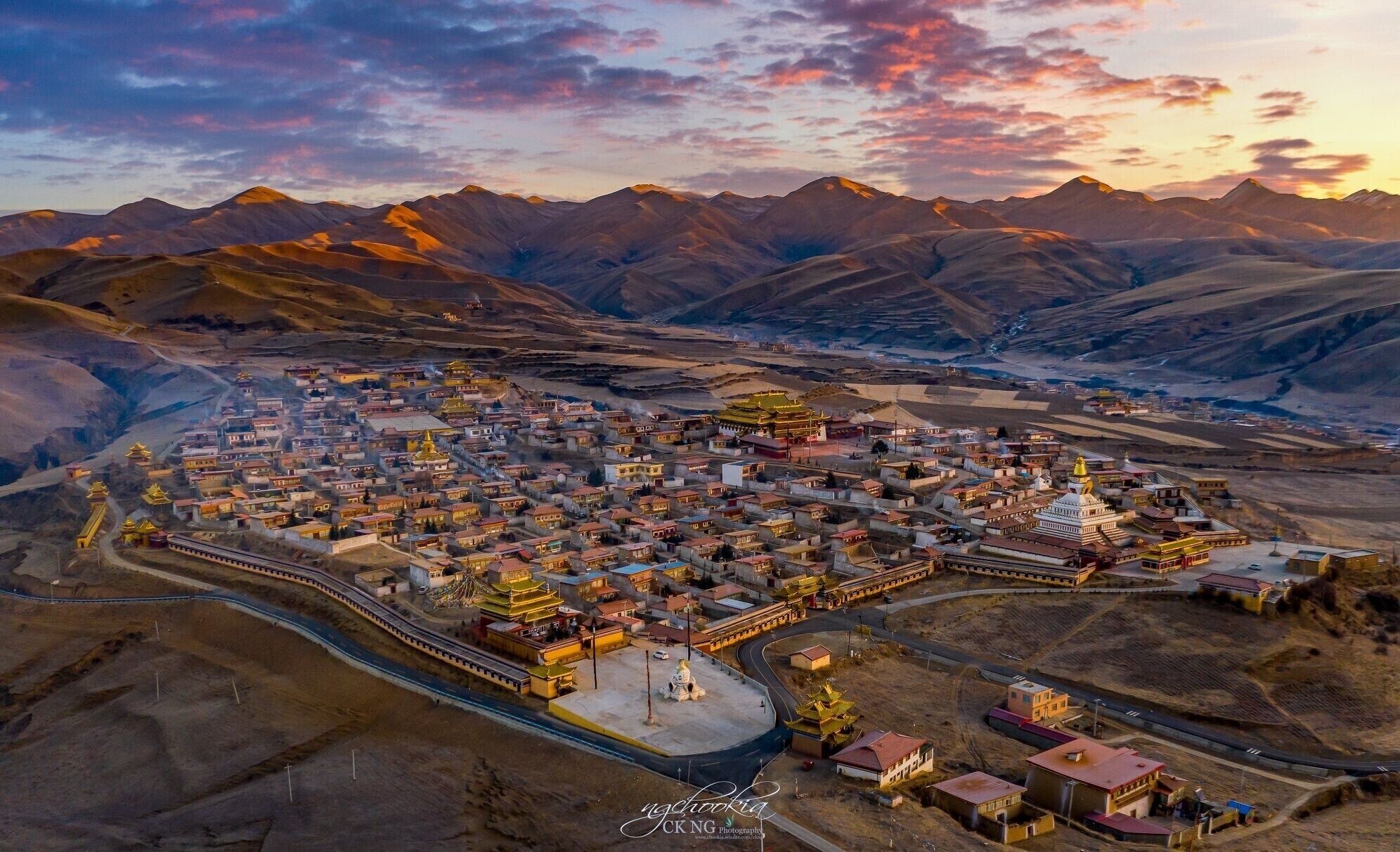 An aerial view of a town with traditional buildings and a prominent golden stupa surrounded by mountains.