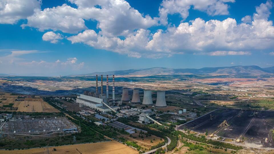 A power plant with cooling towers and multiple chimneys, surrounded by fields and distant mountains.