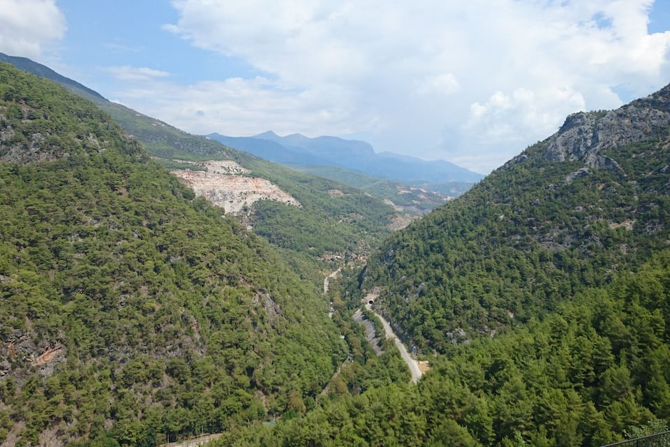Un paysage montagneux avec de vastes forêts verdoyantes, une route sinueuse et des montagnes lointaines sous un ciel partiellement nuageux.