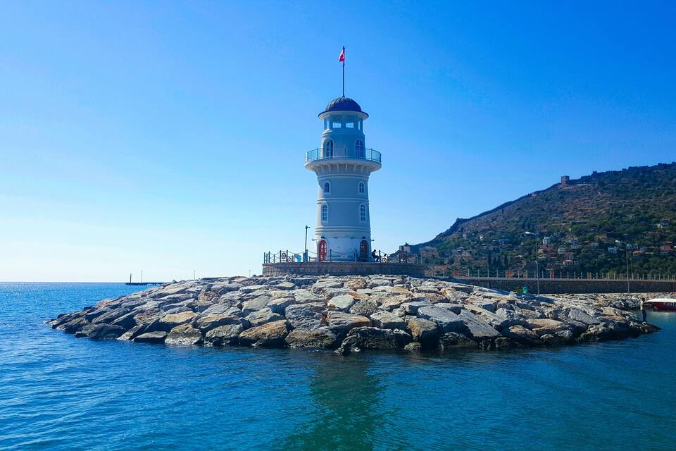 A lighthouse on a rocky outcrop with clear blue water and a hilly backdrop.
