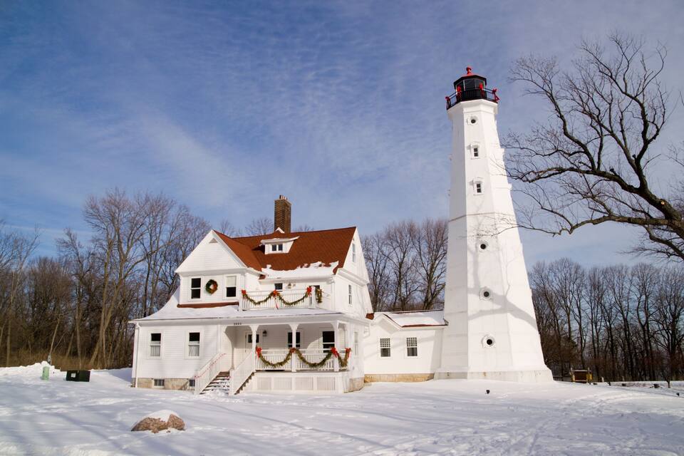 Um farol branco com teto vermelho, uma casa branca com varanda e uma paisagem nevada.
