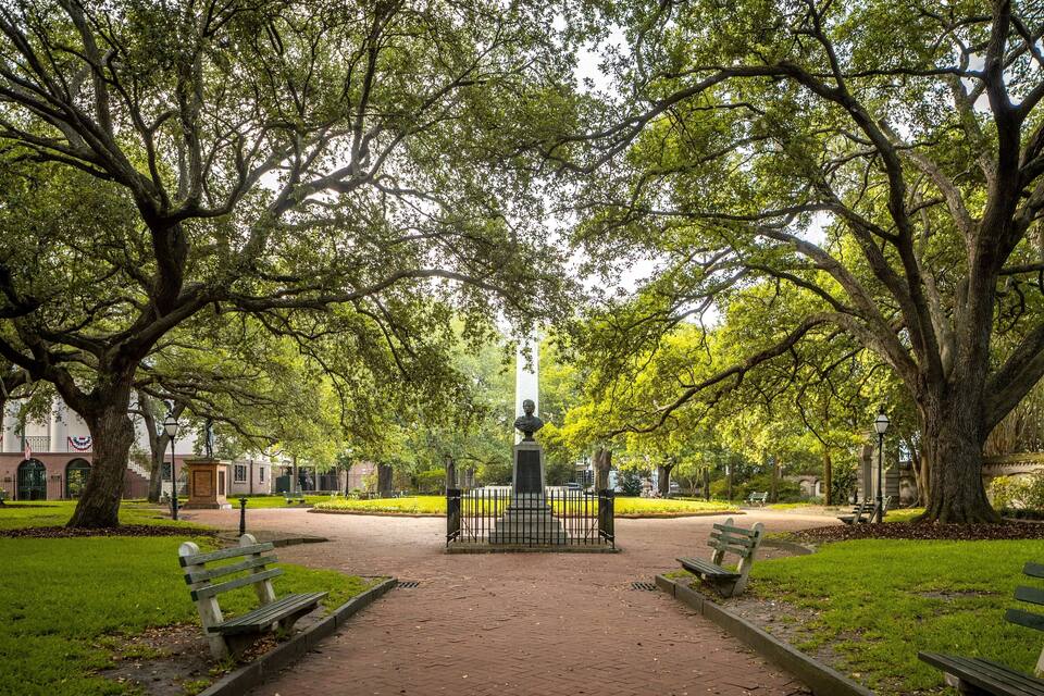 Ein Park mit einem zentralen Denkmal, umgeben von hohen Bäumen und Bänken.