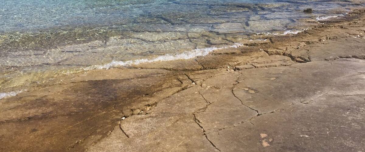 A coastal landscape with clear blue water, rocky shore, and distant mountains.