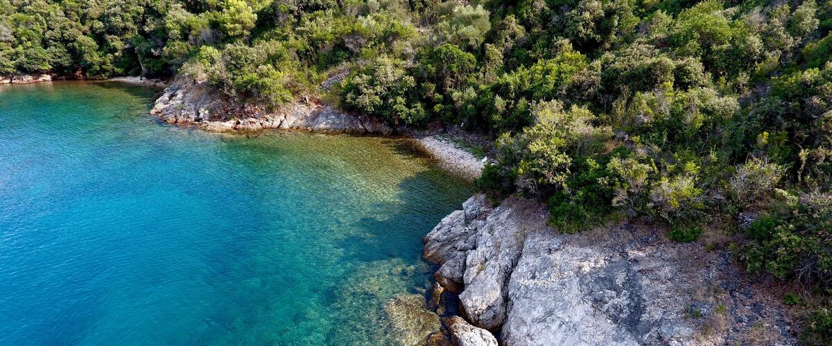 A coastal landscape with clear turquoise waters, rocky shoreline, and dense green vegetation.