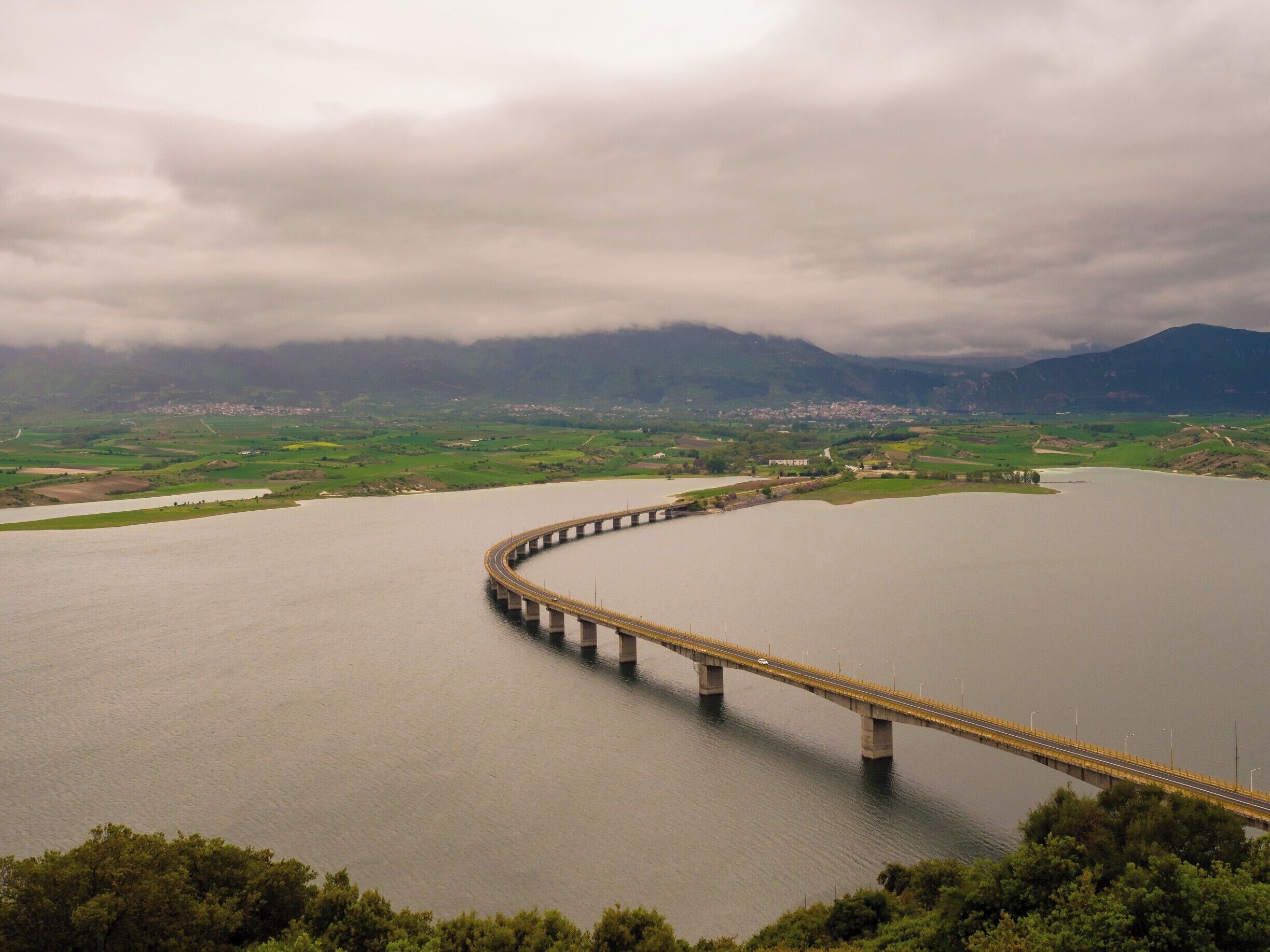 A long bridge spanning a wide river, surrounded by green hills and a cloudy sky.