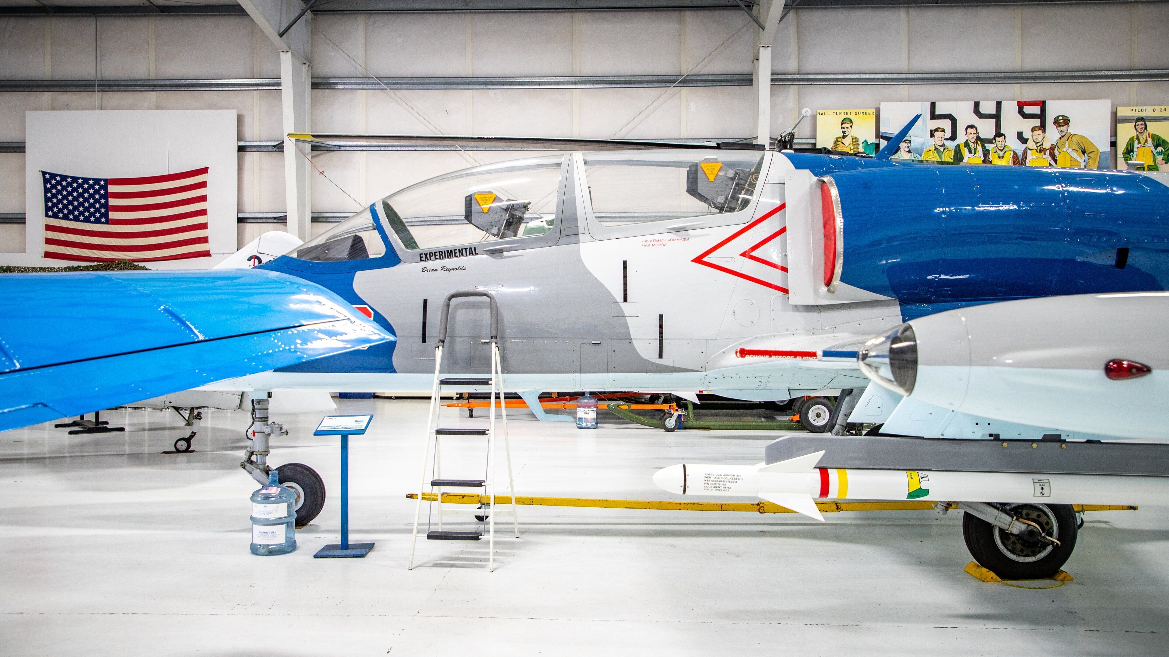 A vintage experimental aircraft with a blue and white color scheme, displayed in a hangar with an American flag and a group portrait in the background.