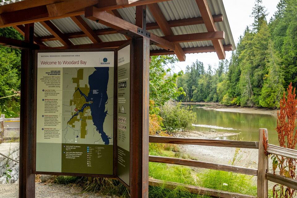 Puinen opastekyltti, jossa on kartta ja tietoa Woodard Bay -lahdesta. Kyltin ympärillä on metsää ja sen vieressä virtaa joki.