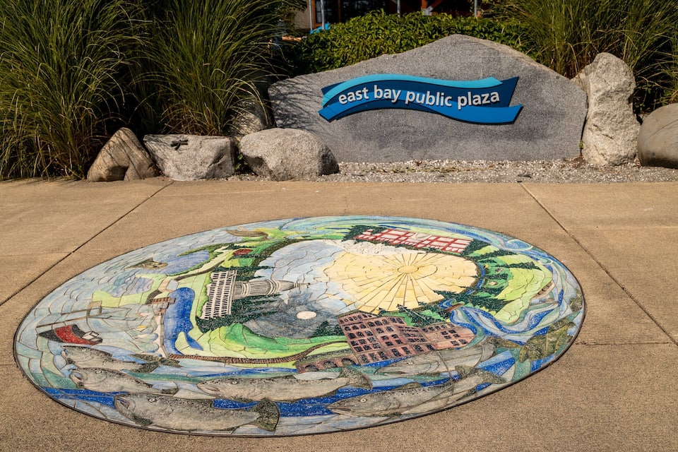 Un mosaic amb peixos i edificis, un rètol que diu "East Bay Public Plaza", i roques i gespa circumdants.