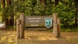 Ein hölzernes Schild mit der Aufschrift 'Millersylvania State Park' und 'Willkommen' in einer bewaldeten Gegend.