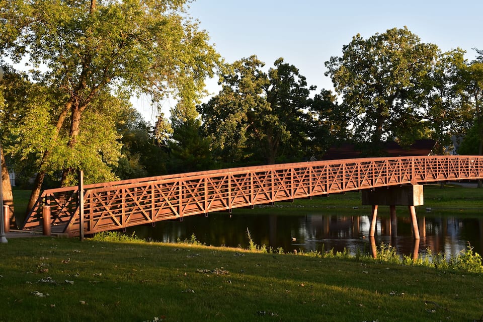 Un puente metálico rojo que cruza un río tranquilo, rodeado de árboles y hierba verdes y frondosas.