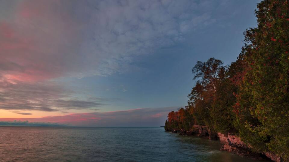 A coastal landscape with a rocky shoreline, dense foliage, and a calm sea under a colorful sky.