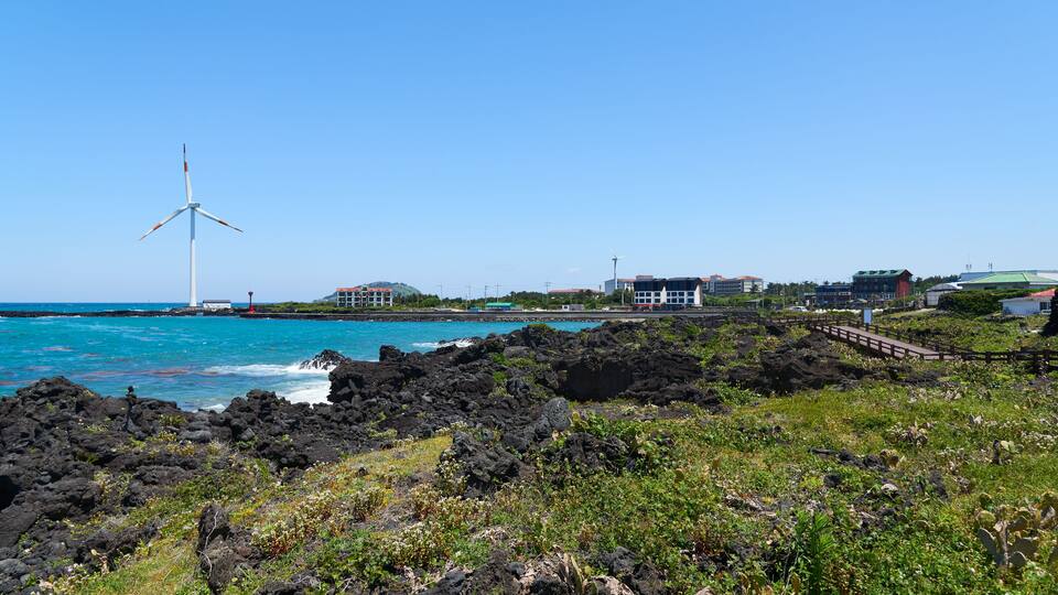 A coastal landscape with a wind turbine, rocky terrain, and buildings along the shore.