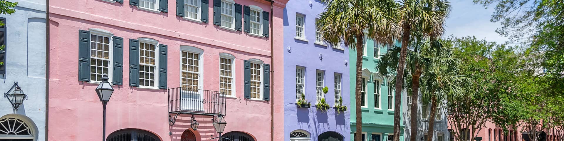 A row of colorful historic buildings with shutters, a street lamp, and palm trees.