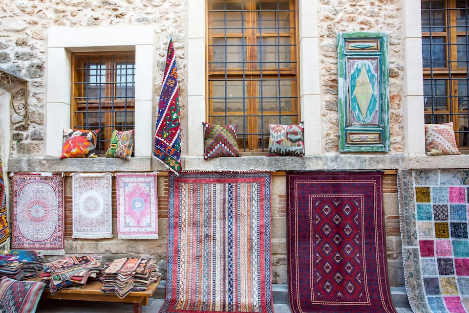 A variety of colorful rugs displayed on a wooden table and hanging on a wall against a stone building with wooden windows.