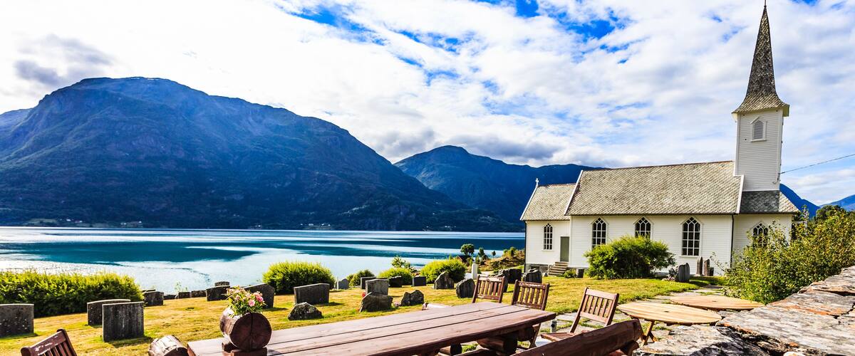 A lakeside church with a wooden bench and chairs, surrounded by mountains and a clear sky.