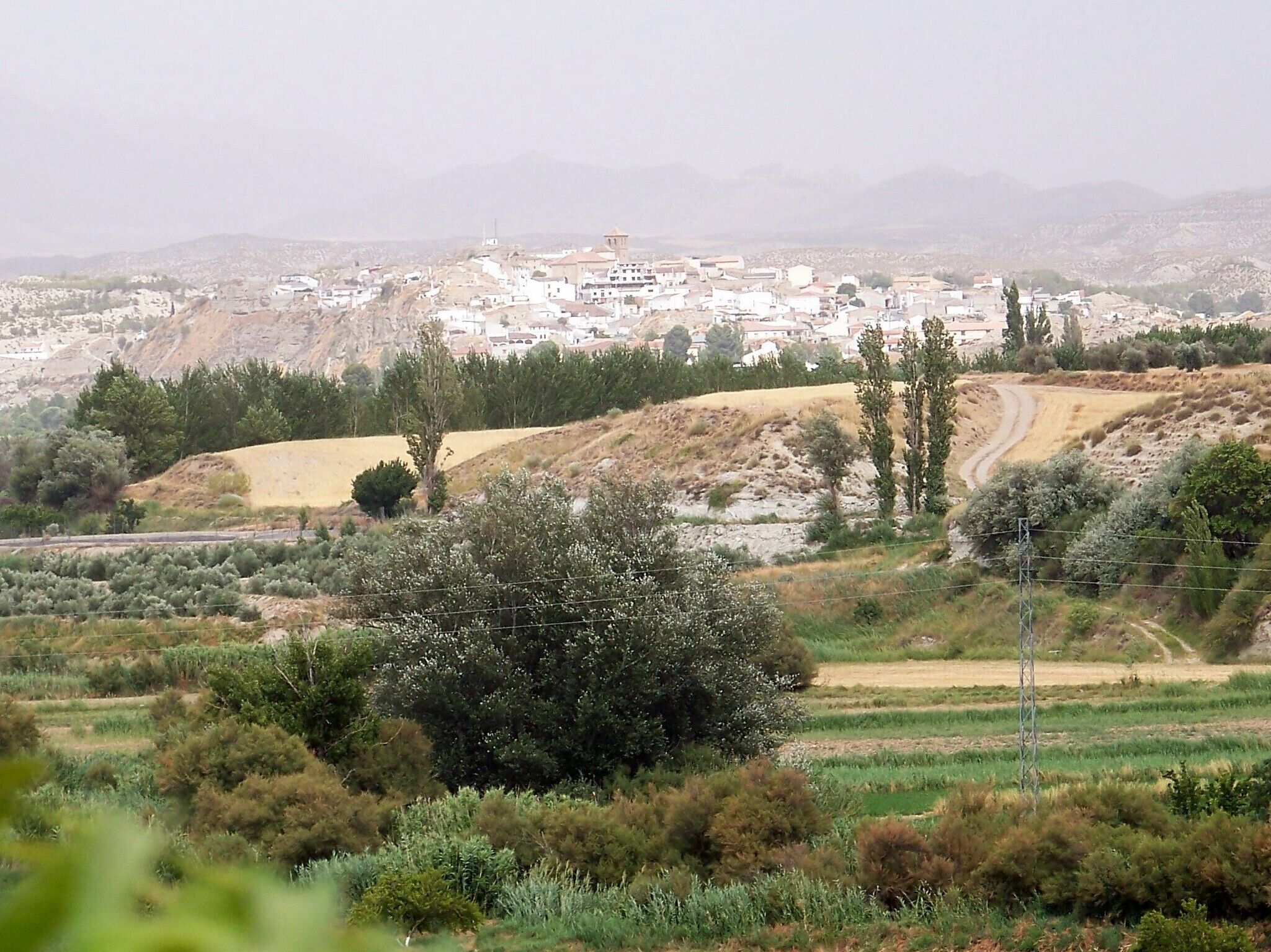 A town with white buildings nestled in a hilly landscape with sparse vegetation and a dirt road.