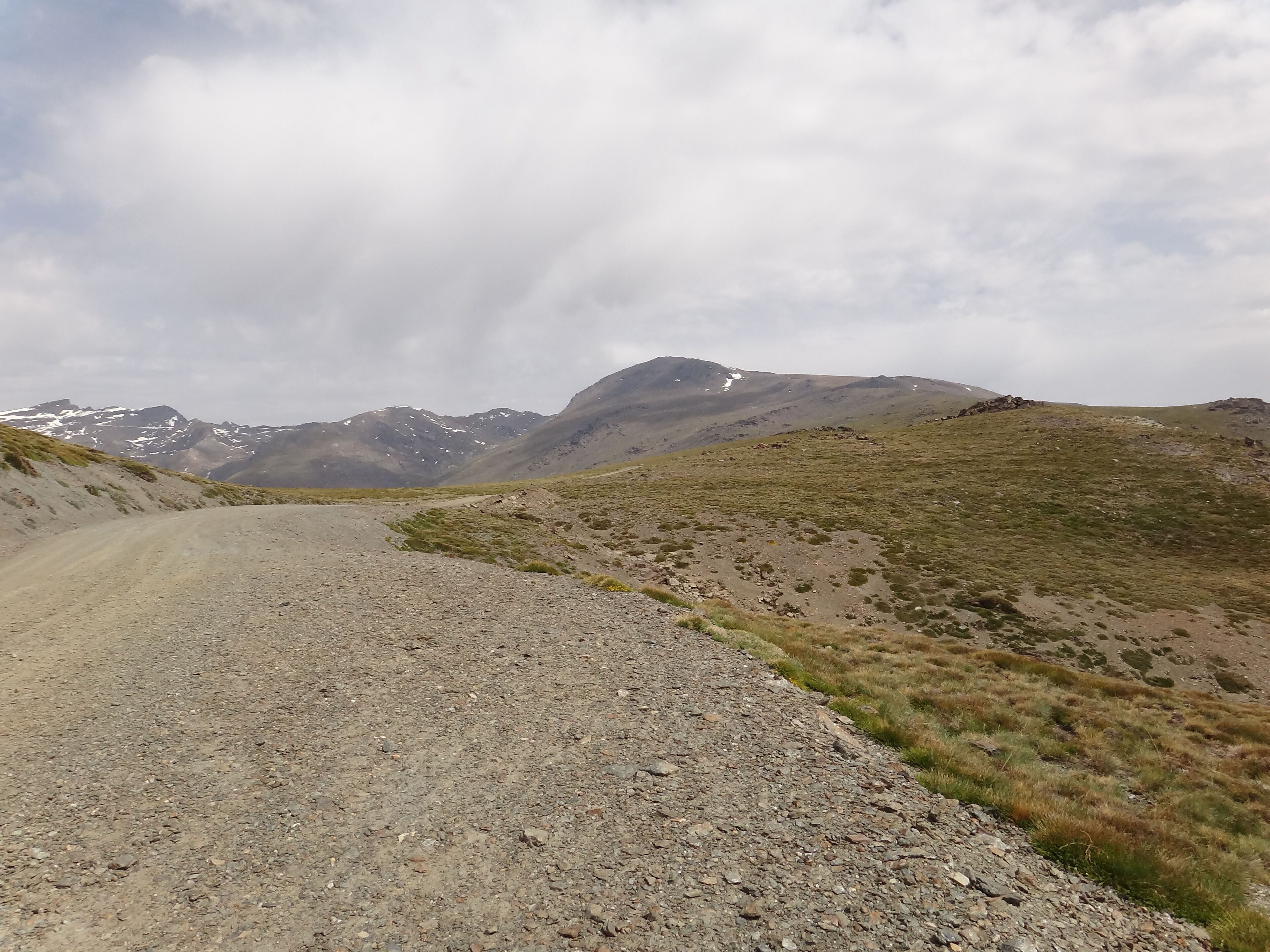 A gravel road winding through a mountainous landscape with sparse vegetation and distant snow-capped peaks.