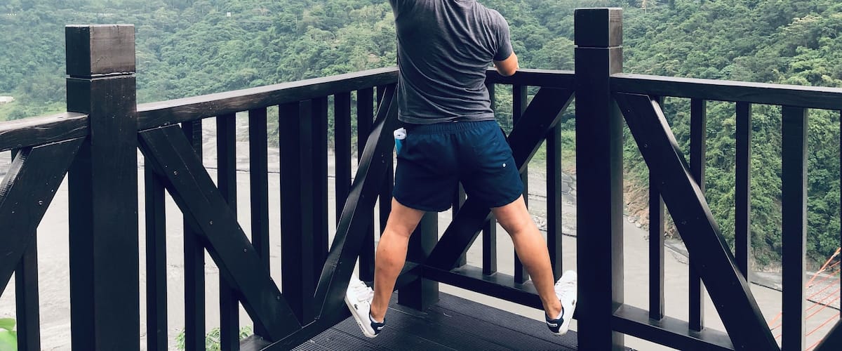 A person standing on a wooden bridge, pointing towards a mountainous landscape.