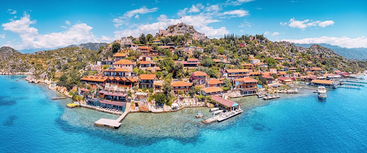 A coastal town with buildings on a hillside, a clear blue sea, and a rocky island in the background.