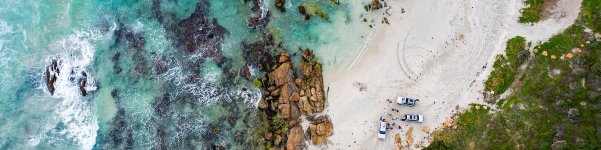 An aerial view of a beach with clear turquoise waters, rocky outcrops, and a few vehicles parked on the sand.