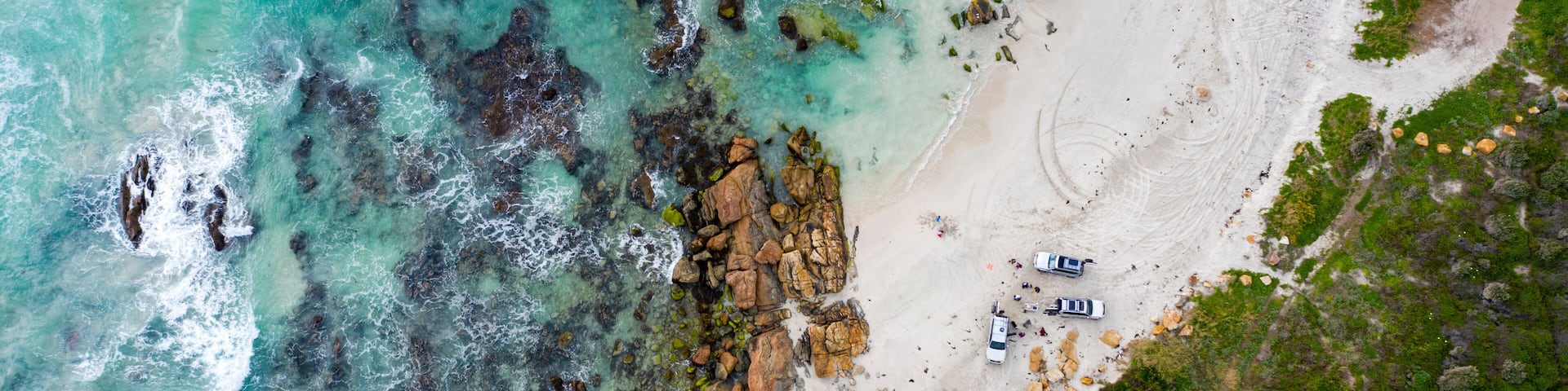 An aerial view of a beach with clear turquoise waters, rocky outcrops, and a few vehicles parked on the sand.