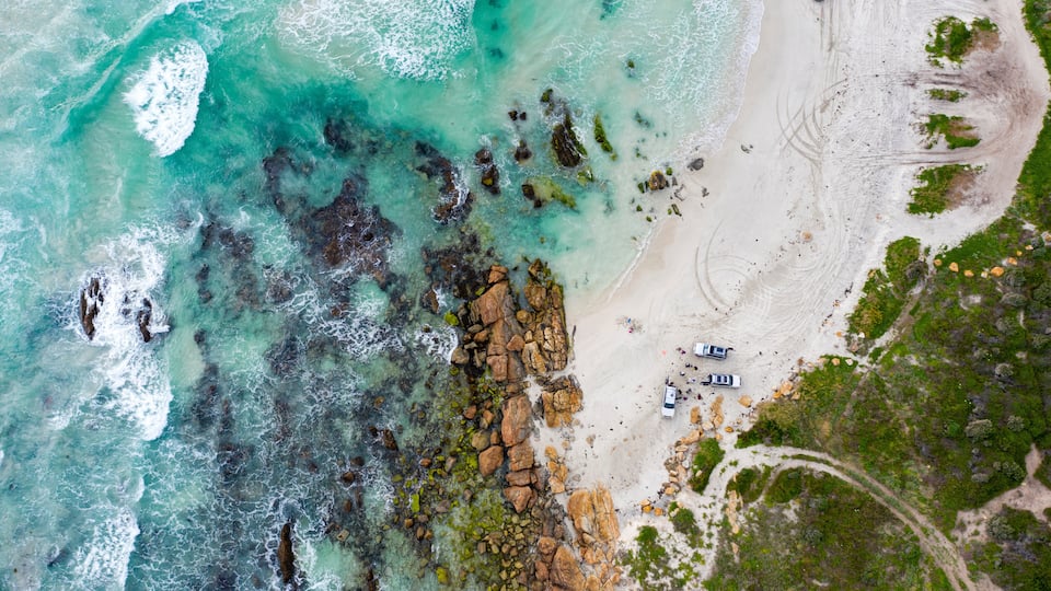 An aerial view of a beach with clear turquoise waters, rocky outcrops, and a few vehicles parked on the sand.