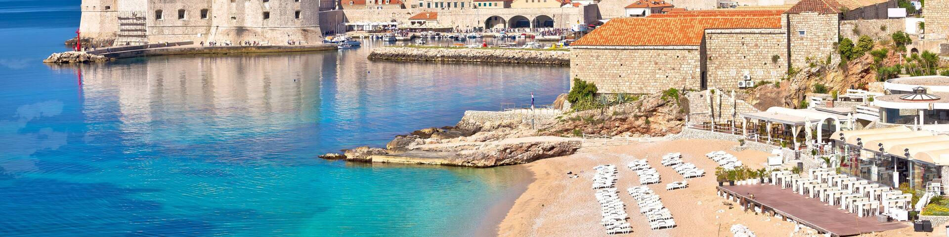 A coastal town with historic buildings, a fortified wall, and a beach with sun loungers.