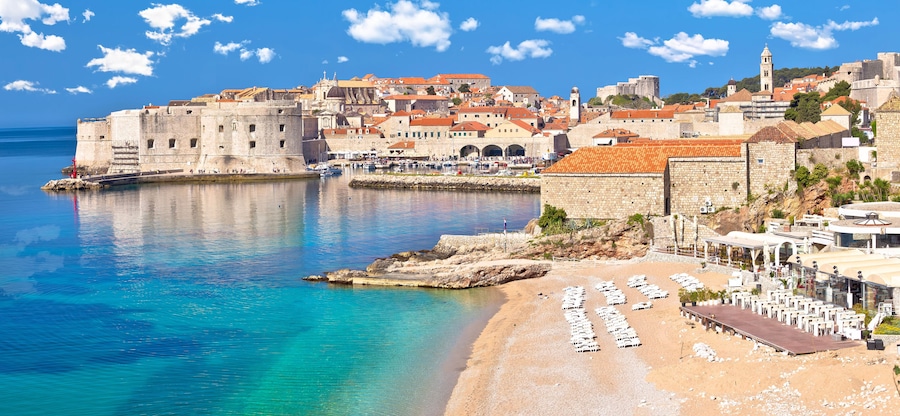 Une ville cĂŽtiĂšre dotĂ©e de bĂątiments historiques, dâune muraille fortifiĂ©e et dâune plage avec des transats.