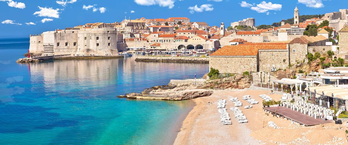 A coastal city with historic buildings, a fortified wall, and a beach with sun loungers.