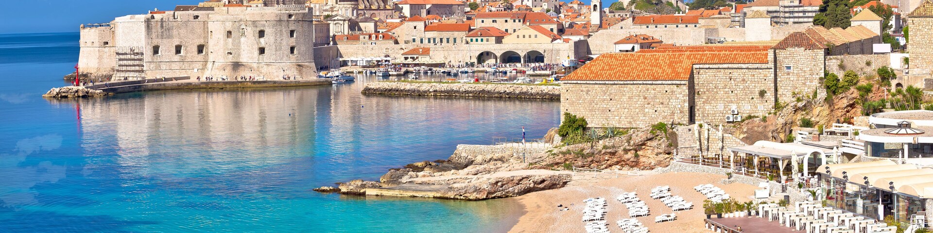 A coastal city with historic buildings, a fortified wall, and a beach with sun loungers.