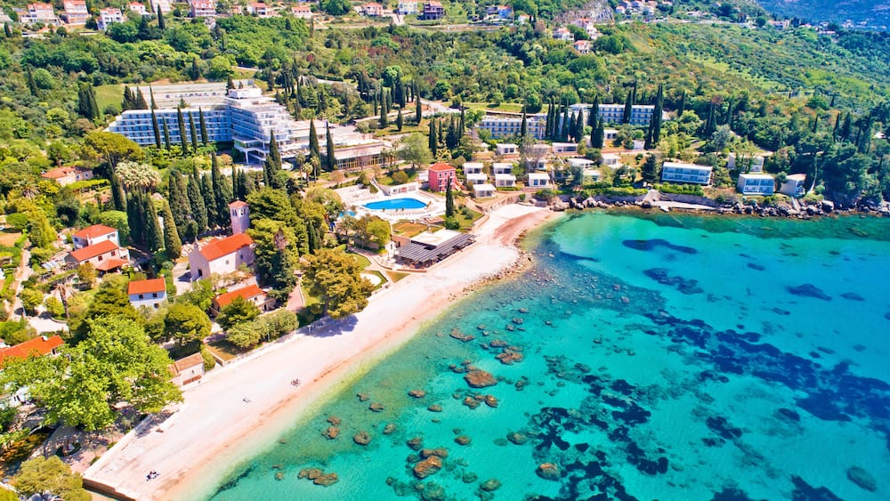 Vue aérienne d’une zone côtière avec une plage, des eaux turquoise et limpides, et un ensemble de bâtiments comprenant un hôtel et des villas.