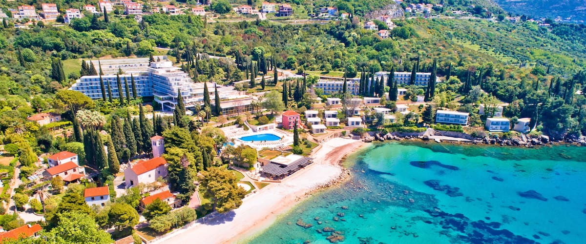 An aerial view of a coastal area with a beach, clear turquoise waters, and a cluster of buildings including a hotel and villas.