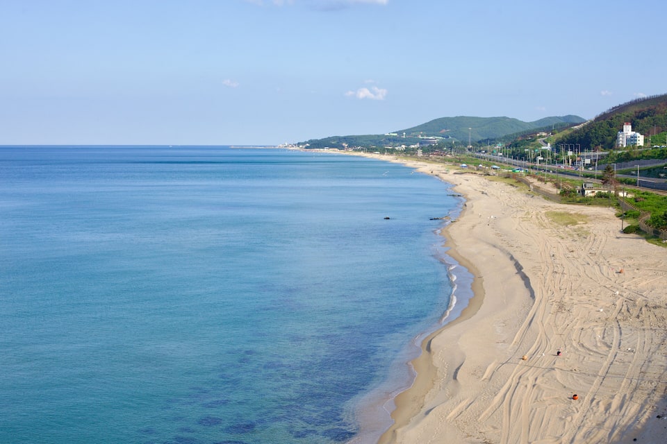 A coastal beach with clear blue water, sandy shore, and a road running parallel to the beach.