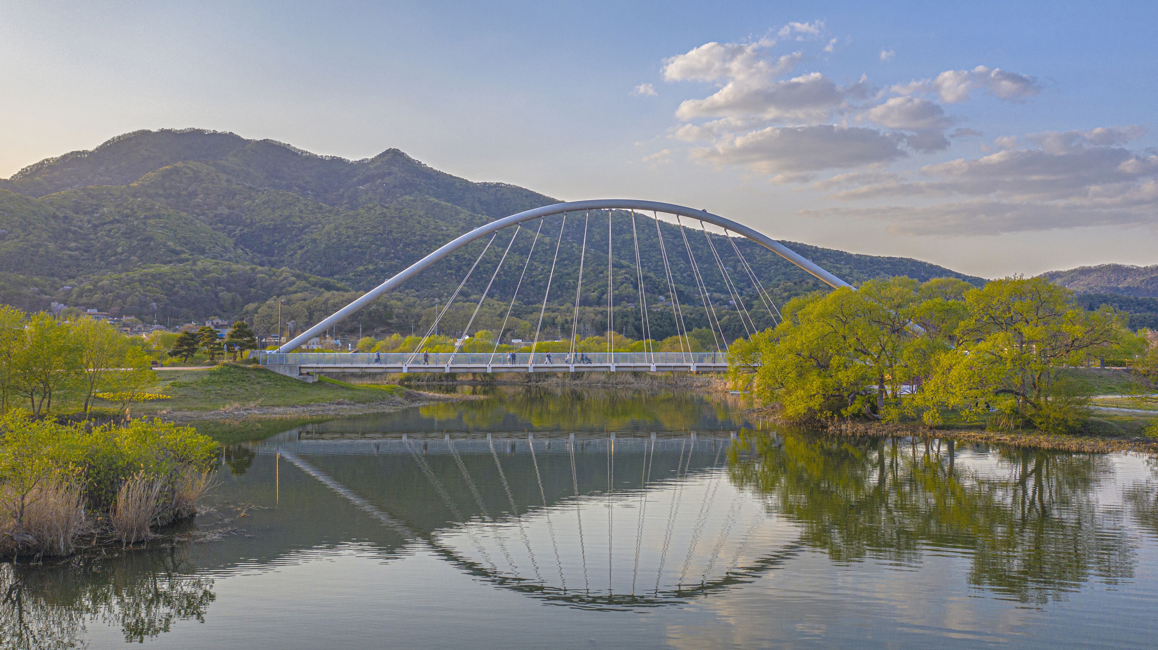A large cable-stayed bridge over a calm river, surrounded by lush green hills and trees.