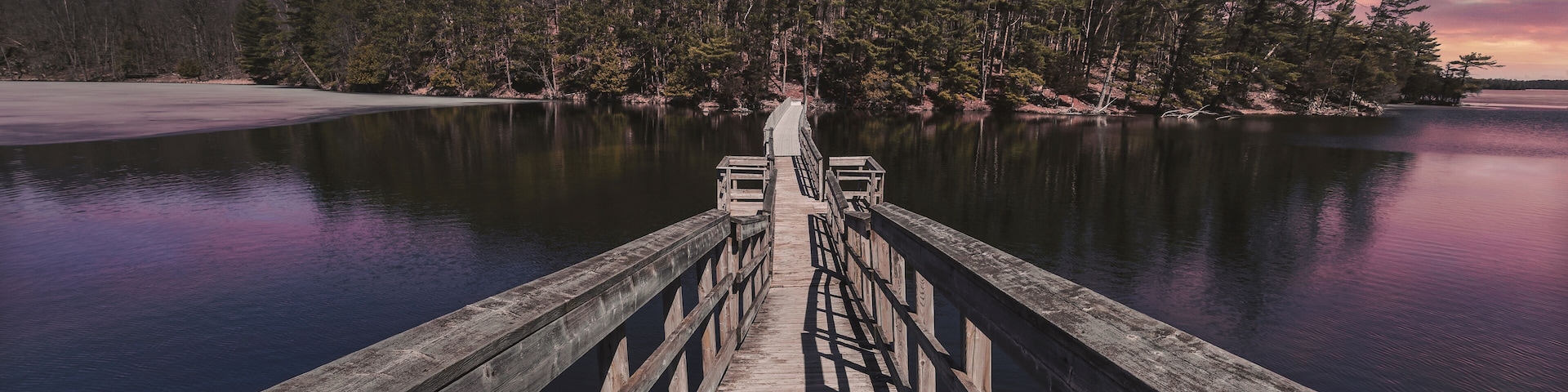 A wooden pier extending into calm waters, surrounded by dense forest and a colorful sky.