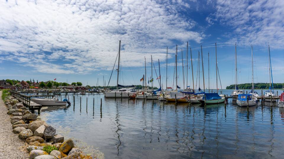 A marina with several sailboats docked, a rocky shoreline, and a clear blue sky with clouds.