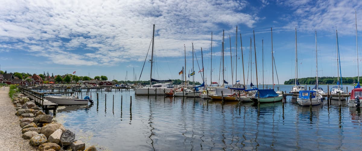 A marina with several sailboats docked, a rocky shoreline, and a clear blue sky with clouds.