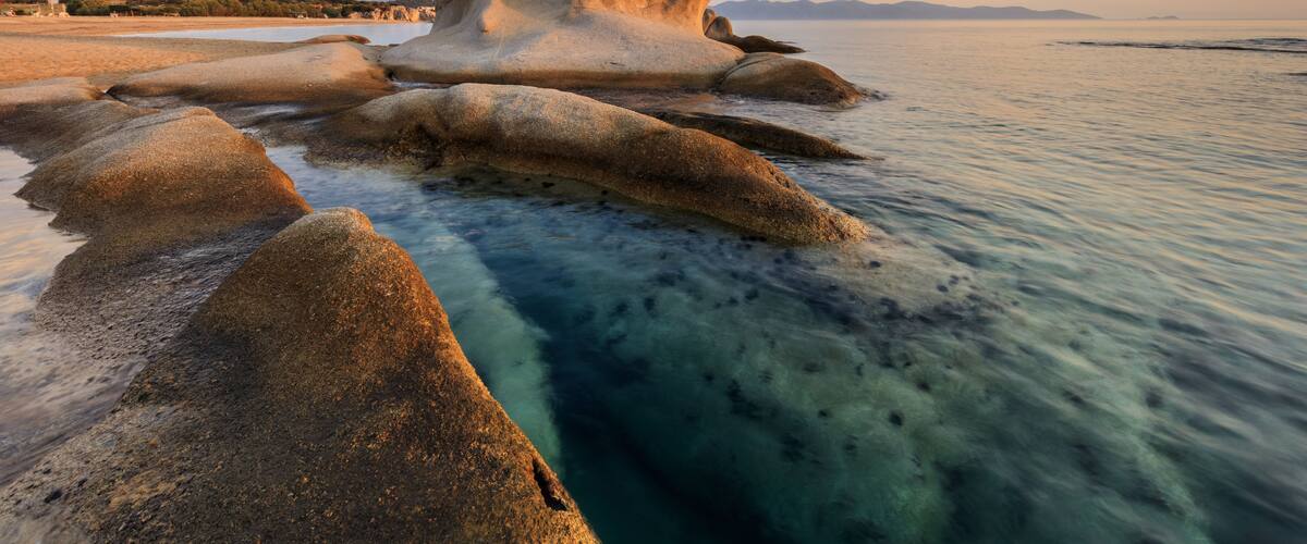 Formación rocosa que emerge de una masa de agua poco profunda, rodeada de agua azul transparente y una costa lejana.