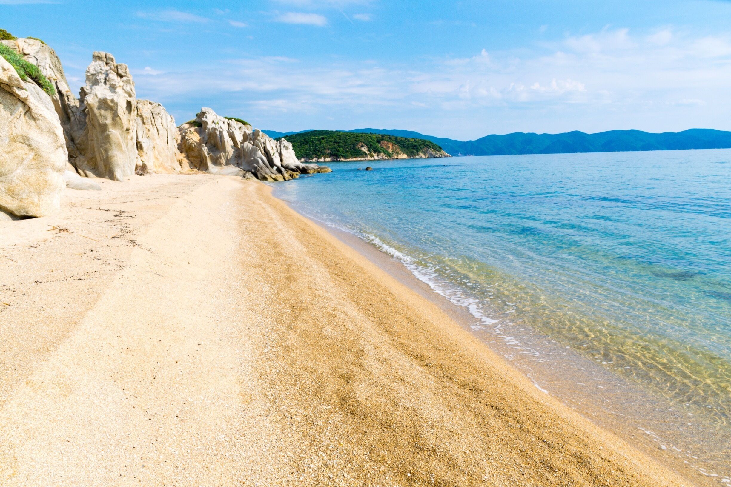 A sandy beach with clear water, rocky cliffs, and green hills in the background.